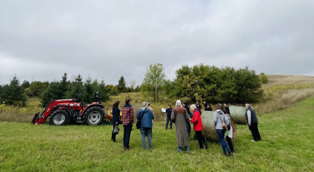 Group of people stand outside in field in front of tractor and hay bales.