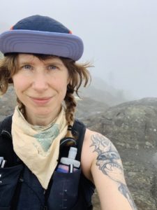 Girl with purple hat and braids stands on foggy, rocky mountain top
