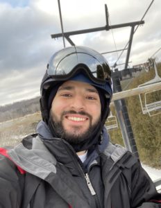 Smiling man in winter jacket, helmet and ski goggles on ski lift with snow dusted landscape in the background.
